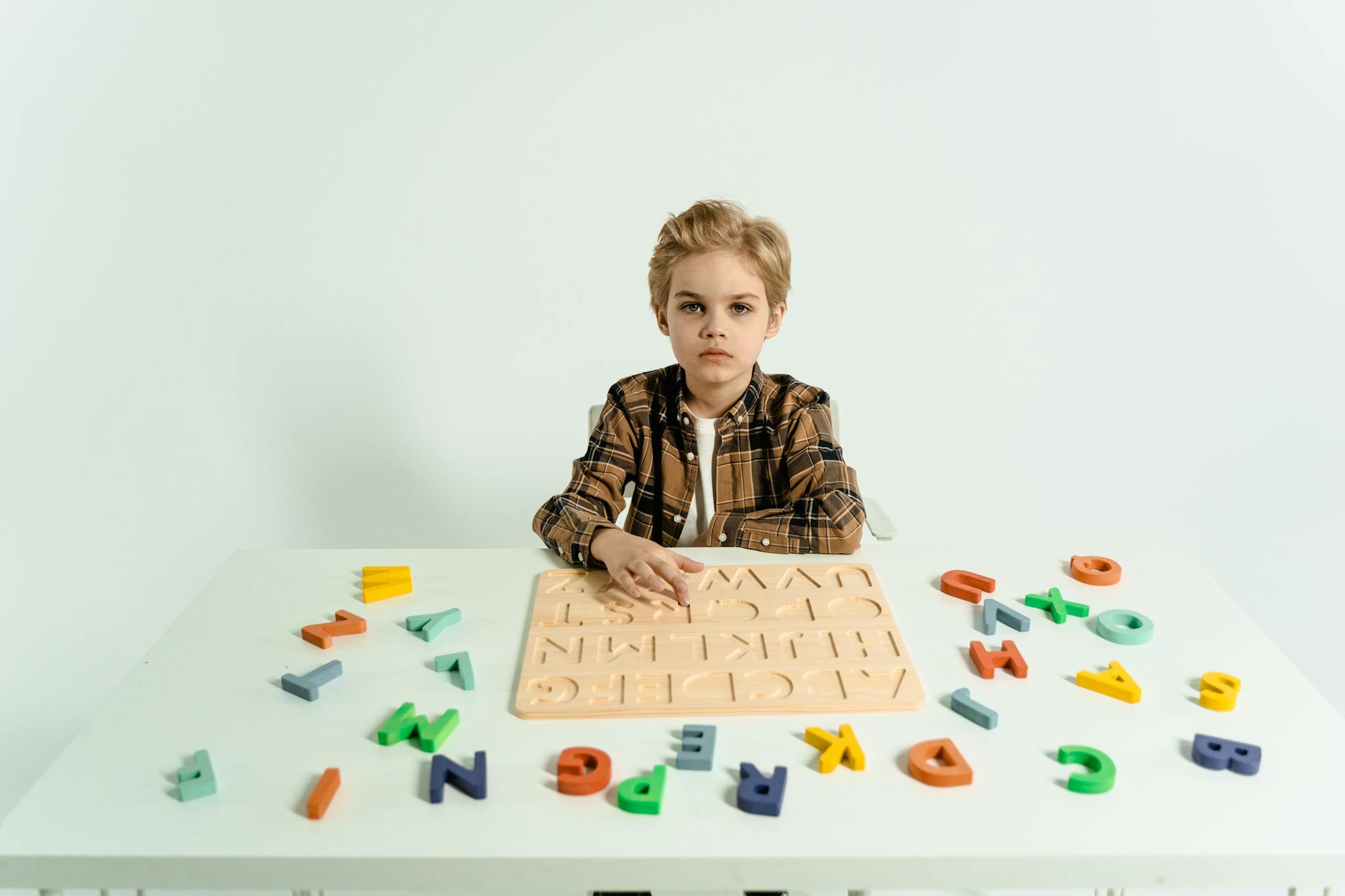 A young boy sits at a table with colorful alphabet puzzle pieces on a white background.
